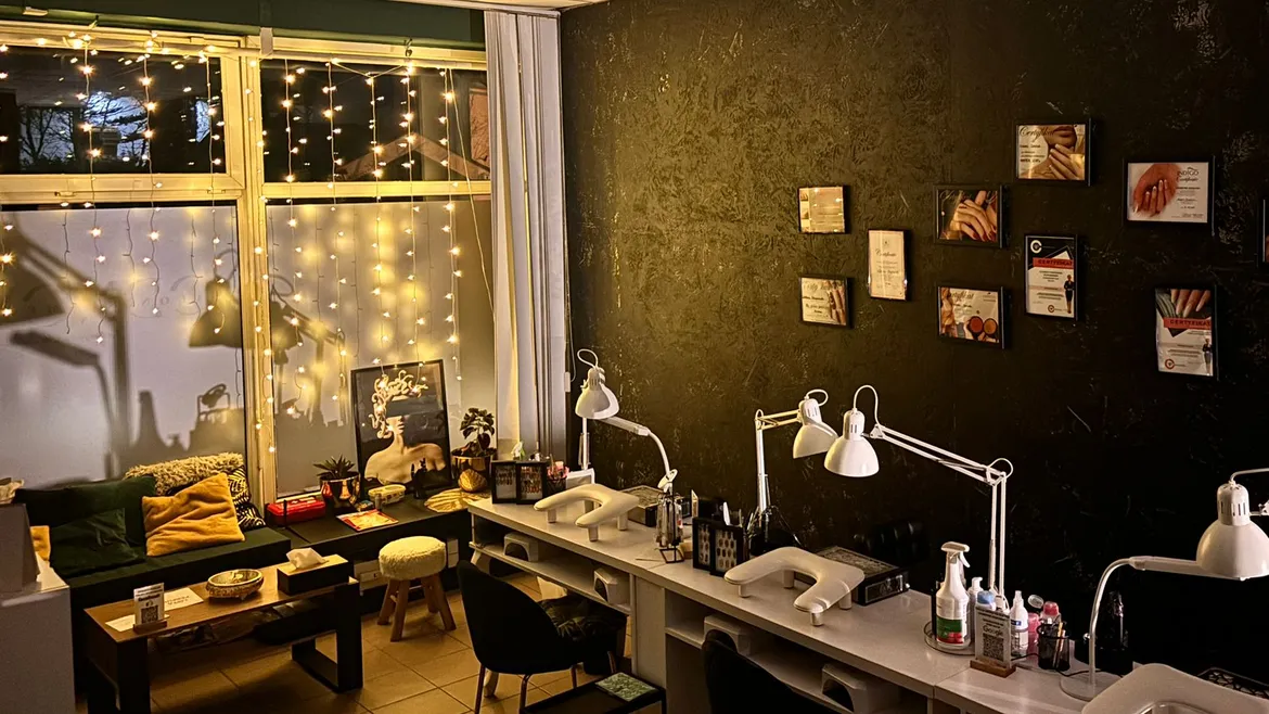 Sala manicure w KAHA wieczorem: czarna ściana z certyfikatami, białe stanowiska z lampami i okno z lampkami
